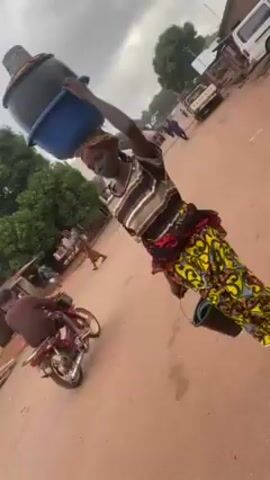 The video depicts a dusty, unpaved road in what appears to be a village or rural area. The sky is overcast. In the initial shot, a woman walks towards the camera carrying a blue bucket on her head and another in her hand. She's wearing a striped shirt, a colorful patterned skirt, and a red wrap around her waist.  As the video continues, the camera follows her movement down the road. Other people are seen walking in the distance, and a few vehicles are parked along the side of the road.  There are small shops or stalls visible along the side of the road as well. As the woman moves further away, two men in casual clothes appear to be talking and gesturing towards the camera.