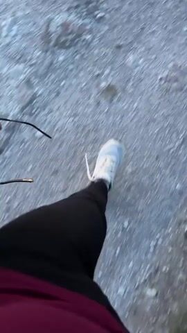 The video shows a first-person perspective of someone walking on a gravel path. The person is wearing white sneakers and dark pants. They are holding a pair of sunglasses in their left hand. The path is surrounded by grass and rolling hills. In the background, the sky is a clear blue with some clouds, and there is a faint sunset visible on the horizon, casting a pinkish hue. The overall environment appears to be a natural, outdoor setting, possibly a mountain or countryside. The video is blurry due to the motion of the camera.
