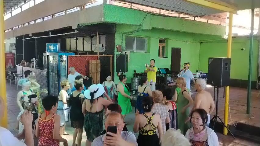 The video shows a group of people, mostly adults, dancing and socializing under a covered outdoor area. The area appears to be some sort of entertainment venue, possibly a bar or restaurant, with a stage or performance area visible in the background. The people are dressed in casual attire, and some are wearing swimwear, suggesting a warm weather setting. Two individuals are standing near a microphone and speakers, possibly as performers or hosts. The lighting indicates it is daytime. The overall atmosphere seems lively and festive.