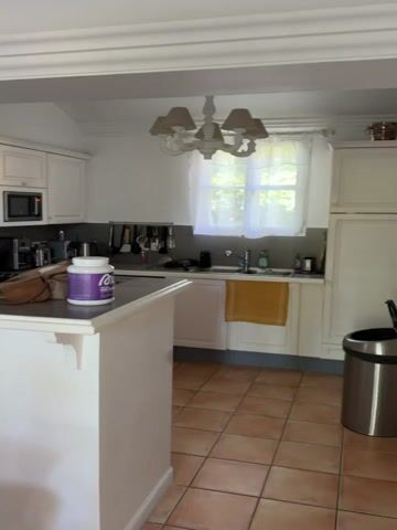 The video shows a kitchen and dining room. The kitchen features white cabinets, a dark countertop, and terracotta tile flooring. The dining room includes a dark wooden table with decorative chairs, a white chandelier, and a black and white framed picture on the wall. A white rocking horse and a water bottle are on the table. A pair of white French doors lead to an outdoor patio with a wooden deck, a table with chairs, and a lavender umbrella. The camera pans from the kitchen to the dining room and then focuses on the French doors and the view of the patio.