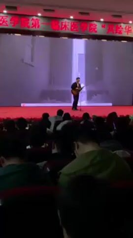 A person is standing on a red stage in front of an audience, playing an acoustic guitar and singing into a microphone. A large screen behind the person displays an animation of a white paper airplane flying through a corridor. The audience is seated in rows of red chairs, most are looking at their phones. There is Chinese text at the top of the screen above the stage. The room appears to be an auditorium or lecture hall. The lighting is primarily provided by red overhead lights.