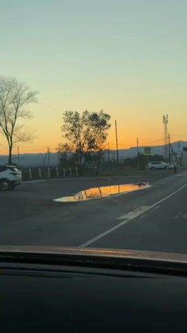 The video shows a view from inside a vehicle looking out at a rural road during sunset. In the foreground, the dashboard of the car is visible. The road has a large puddle reflecting the orange and yellow hues of the sky. On the side of the road, there is a tree, a pedestrian crossing sign, and a traffic sign. Further down the road, there is a white car parked, and two people are standing near the crosswalk. A cell phone tower and some buildings are visible in the background. The sky is a clear gradient of pale blue at the top transitioning to orange and yellow near the horizon, indicating the sun is setting.