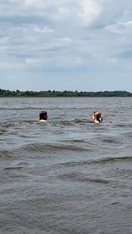 Two people swim in a body of water. The person on the left has dark hair and appears to be swimming on their back. The person on the right has lighter hair and is swimming on their stomach, with their head above water. They are in a large body of water with waves. The background shows a green tree line and a cloudy sky.