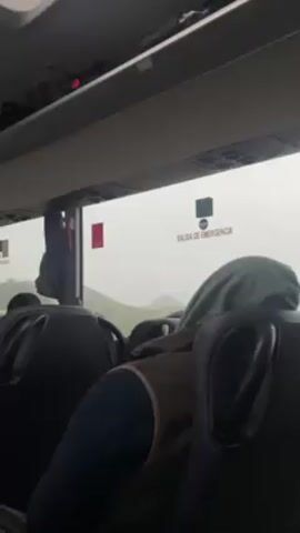 The video shows the interior of a bus, with the focus on the back of a passenger's head resting against the seat. The passenger is wearing a dark blue shirt and a brown vest, with a green hood pulled over their head. The window shows a foggy or misty view of what appears to be mountains. There is an emergency exit sign in Spanish above the window that reads "SALIDA DE EMERGENCIA." Luggage racks are visible above the seats. The bus appears to be in motion.
