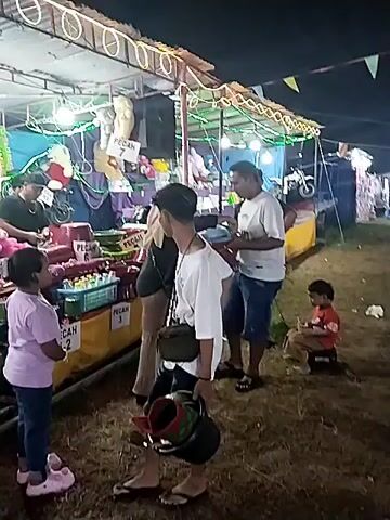 The video shows a nighttime scene at what appears to be a fair or carnival. In the foreground, several people are gathered around a game booth. Two men are filling buckets with what look like small prizes or toys. A young girl in a pink shirt and sandals stands near the booth, watching. Another young child sits on a small stool, also watching. The game booth is decorated with lights and has prizes displayed, including plush toys. A sign on the booth reads "PECAH 1-2." Other people are visible behind the booth, seemingly running the game. The ground is grassy and littered with debris. The overall atmosphere suggests a casual, festive environment.