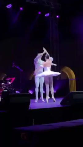 Two ballet dancers, a man and a woman, are performing on a stage. They are both dressed in white costumes, with the woman wearing a tutu and tiara. The man is holding the woman's hand and guiding her through a series of ballet moves. They are in front of a backdrop that is mostly dark. There are stage lights above them, and a drum set is to the left of the stage. The lighting is purple, and speaker equipment sits at the bottom of the screen.