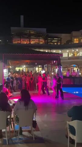 The video opens with a wide shot of an outdoor nighttime event. People are gathered around a lit dance floor, where a couple is performing a dance routine. The dance floor is illuminated by pink and purple lights. In the background, there is a building with multiple levels, likely a hotel or resort, with lights visible inside. A swimming pool is visible to the right of the dance floor. The camera zooms in on the dancers as the song progresses. The dancers are dressed in performance attire. The overall atmosphere appears to be festive and social, with an audience watching the performance.