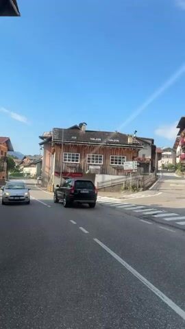 The video shows a street in a European town on a sunny day. Several cars are driving on the road, including a gray SUV, a black SUV, a silver hatchback, and a blue hatchback. A pedestrian is visible crossing the street at a crosswalk. The buildings lining the street are colorful and have a traditional European architectural style, with balconies adorned with flowers. The sky is clear and blue with a few wispy clouds. The overall impression is one of a quiet, pleasant town with moderate traffic.