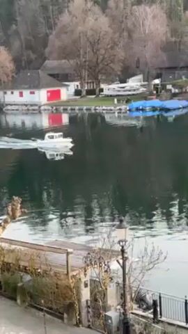 The video shows a white motorboat moving on a lake towards the right side of the frame. On the shore, there's a building with a red section and several boats covered with blue tarps. Other buildings and trees line the shore as well. The foreground includes a decorative outdoor area with a lamp post. The overall lighting suggests a slightly overcast day.