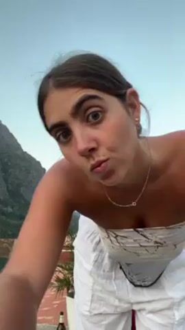 The video shows a woman standing on a patio or balcony with a mountain in the background. The sky is blue, and it appears to be early evening. She is wearing a white scarf tied as a strapless top and white linen pants. The camera is positioned at a low angle, looking up at her. She poses briefly, looking at the camera. There is a wooden railing, a red rooftop, and some vegetation visible in the background. A bottle is seen on the patio.