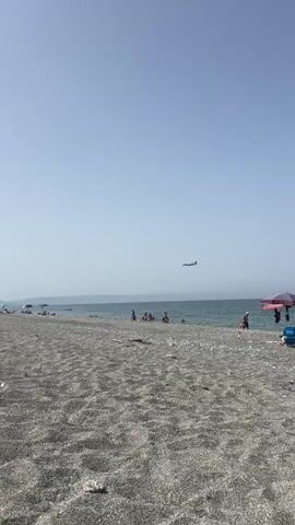 The video shows a wide shot of a pebble beach on a clear, sunny day. The sky is a pale blue, and the sea is a muted blue-green. The beach is covered in small, light-colored pebbles, with some larger pieces of driftwood scattered about. In the distance, the shoreline is visible with a line of hazy mountains. Several people are present on the beach and in the water. On the beach, some are sitting or lying under umbrellas, while others are standing or walking. A small group of people, including children, are in the shallow water. To the right of the frame, a man in red shorts is walking along the water's edge. The most prominent feature is an airplane flying horizontally across the upper portion of the frame. The plane is small in the distance and appears to be on a descent or approach. The overall atmosphere is one of a relaxed summer day at the beach.