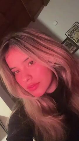 A young woman with long, blonde hair is shown in a close-up shot. She is looking directly at the camera. The lighting is red, casting a warm glow on her face. She is wearing a black top. In the background, there is a wall with two framed pictures.