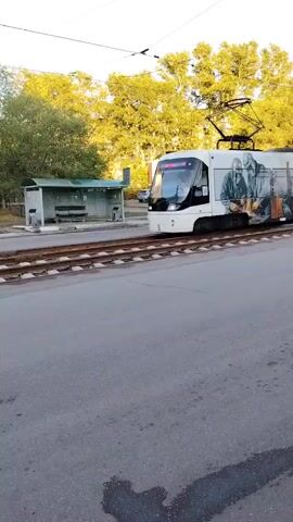 This media file captures a tram with a distinctive mural on its side, moving along tracks. The mural depicts several individuals, seemingly workers, wearing protective gear including helmets and masks, and engaged in an activity that appears to be welding, indicated by the sparks and torches. The scene is set outdoors, with a clear sky and dappled sunlight suggesting it's daytime, likely morning or late afternoon given the angle of the sun.

Behind the tram, there are trees with green foliage, and a modest building with several windows, possibly an apartment block or administrative facility. To the left, a small, simple shelter with a bench is visible, likely a bus stop or tram stop. The tram itself is white with the mural covering a significant portion of its length. The tram bears the number "8" on its front. The overall setting appears to be urban or suburban, with a road in the foreground and the tram tracks running parallel to it. The weather seems fair and pleasant. The mood is neutral, focusing on the visual elements of the tram and its surroundings. No discernible text other than the number "8" is present. The location context provided is Temirtau, Kazakhstan.