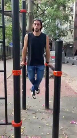 The video features a man doing dips on parallel bars in an outdoor setting. He is wearing a black sleeveless shirt, jeans, and black and white sneakers. The parallel bars are black with orange connectors, situated on a playground or exercise area. A tree is visible in the background, along with typical urban elements such as buildings, sidewalks, and street signs. The ground beneath the bars is covered with fallen leaves and flower petals. The man demonstrates the dip exercise by lowering his body between the bars and then pushing himself back up, repeating the motion several times. The scene is recorded at eye level in daylight.