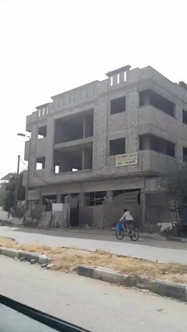 The video shows a gray, overcast day as seen from a moving vehicle on a road. Buildings with unfinished construction are visible on the side of the road.  One building has multiple levels, with hollow window spaces and a plain facade.  A second building with a similar unfinished structure is shown at a distance. A cyclist wearing a blue shirt is seen in the first view, and there are some tattered cloth coverings in front of the first building. The road has a concrete curb. A couple of electrical poles with power lines are visible. There is no dialogue or other audio information.