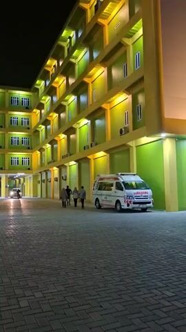 This video captures the exterior of a brightly lit building at night, likely a hospital or medical facility, in Lampeuneurut Blang, Indonesia. The building features a distinctive facade with alternating bands of green and yellow. Numerous windows, possibly air conditioning units, and architectural details are illuminated by yellow lighting, creating a vibrant and welcoming atmosphere despite the darkness.

In the foreground, a paved courtyard with a distinctive brick pattern is visible. Several people are walking across the courtyard, suggesting some level of activity at the facility. An ambulance, marked with red and white accents, is parked on the right side of the frame, indicating the presence of emergency services. In the background, a car is seen driving through an open entryway or a covered walkway of the building.

The overall scene is well-lit, indicating the building is operational. The presence of an ambulance and the signage "IGD 24 JAM" (which translates to Emergency Department 24 Hours) confirm this is a medical emergency facility. The time of day is clearly night, with the dark sky visible above the illuminated building. There are no obvious indications of weather conditions other than the clear night sky. The emotions evoked by the scene are a mix of urgency, due to the ambulance, and a sense of readiness and accessibility, highlighted by the continuous operation indicated by the signage and the bright illumination.