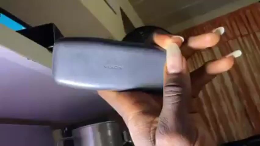 A person with long fingernails is shown holding a gray Nokia feature phone. The phone is being held in a way that displays both the back of the phone with the Nokia logo, and then the keypad on the front. The background appears to be a kitchen or pantry setting with cabinets and curtains. There is a stainless steel pot or container visible below the phone. The camera angle is from below, looking up at the person's hand and the phone. The lighting is mixed, with some areas appearing darker and others brighter.