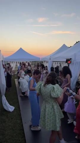 A group of people, predominantly women and a few children, are dancing outdoors on a dark wooden dance floor at sunset. They are holding hands and forming a line, with some dancing in a circle. In the foreground, a woman in a light green patterned dress with puff sleeves dances with her back to the camera, holding hands with a woman in a blue jumpsuit. Further down the line, a woman in a white gown is dancing and smiling. Several white pointed tents with draped fabric decorations are visible in the background, set against an orange, pink, and blue sky. Other guests are standing around the dance floor on the grass, watching the dancers.