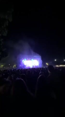 The video shows a concert at night. The stage is brightly lit with white and blue lights. There is a large screen behind the band displaying the band's name, Nine Mile, and an image of one of the members. The band is performing on stage, with a singer in the center and other members playing instruments around him. A large crowd of people is watching the performance, their silhouettes visible in the foreground. The atmosphere is energetic, with music and flashing lights.