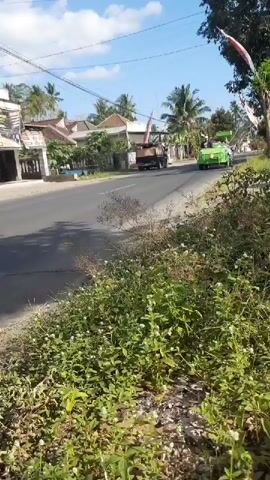 A green vehicle, possibly a modified tractor or a utility vehicle, is traveling on a paved road. The vehicle has a person driving it, and it appears to be carrying some sort of equipment or cargo on the back. The road is lined with vegetation, including bushes and grass, on the right side. In the background, there are buildings that resemble houses or small businesses, and palm trees. The sky is blue with some clouds. Another vehicle, possibly a truck, is visible ahead of the green vehicle. The overall scene suggests a rural or semi-urban environment on a sunny day.