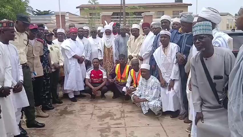 A large group of people, predominantly men, are gathered outdoors in what appears to be a semi-urban or suburban setting in Sabon Gari North, Nigeria. The group is diverse in age and attire. Many men are wearing traditional Nigerian clothing, including flowing white robes (baba riga) and various types of caps and turbans, some of which are elaborately decorated. Several individuals are in military or security uniforms. Three men in the foreground are kneeling or crouching, one wearing a red and white soccer jersey with "Emirates" and "flybetter" visible, another in a dark outfit, and a third in a checkered robe. Two men in the center of the group are wearing bright orange safety vests over their clothing.

The group appears to be posing for a photograph or video, with many looking towards the camera. The overall mood seems formal and possibly ceremonial, given the attire of many attendees. The background shows a building with light-colored walls and some vegetation. The lighting suggests it is daytime, likely late morning or early afternoon, with overcast or diffused sunlight. There are no strong shadows visible. The ground is paved with reddish-brown tiles or bricks.

Notable details include the specific clothing worn by the individuals, the presence of uniformed personnel, and the gathering of a large number of people in a public or semi-public space. The "Emirates" and "flybetter" text on the soccer jersey provides a specific brand detail. The context of Sabon Gari North, Nigeria, indicates a likely cultural and geographical setting for this event. The scene captures a moment of collective gathering and potentially an organized event or meeting.
