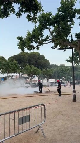 The video captures a scene in a public park, likely on a beach or waterfront, given the sandy ground and trees. Two individuals in dark uniforms and red helmets, appearing to be emergency responders or civil protection personnel, are actively engaged in extinguishing a small fire. One person is holding a hose, directing a powerful stream of white foam or water onto the smoldering area, which is producing a significant amount of white smoke. The other person stands nearby, holding a hose, ready to assist. In the background, several parked cars are visible, along with more trees and buildings suggesting an urban or semi-urban environment. Further in the distance, a few other people can be seen, seemingly observing the situation. The scene appears to be taking place during daylight, possibly late afternoon or early evening, with the sky a pale blue. The overall mood is one of controlled action and public safety. Visible text on the uniforms of the responders reads "PROTECCIÓ CIVIL" and "AJUNTAMENT DE GUÍXOLS", indicating their affiliation with Civil Protection and the municipality of Guíxols, Spain. Red and white striped caution tape is set up in the foreground, cordoning off the area and further emphasizing the presence of an incident.