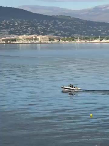 The video shows a calm, blue bay on a sunny day. A small, white motorboat is moving across the water, leaving a wake behind it. Two people are visible in the boat. In the distance, a coastline with buildings and a mountain range can be seen. A single yellow buoy floats near the bottom right corner of the frame.