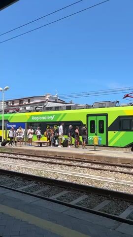 A bright green and yellow train with "Regionale" and "Tropea" written on its side is stopped at a train station platform. Several people, some with luggage, are standing on the platform near the train doors. One person is boarding the train, and another appears to be waiting. The sky is clear and blue. The train tracks are visible in the foreground.