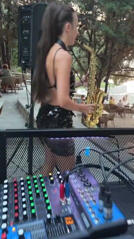 The video shows a woman playing a saxophone outdoors. She is wearing a black sequined dress with an open back and her hair is pulled up in a high ponytail. The saxophone is a golden tenor saxophone. In the foreground, a DJ mixing board with illuminated green and red lights is visible. In the background, there are tables and chairs suggesting a restaurant or outdoor seating area, with trees and foliage forming a natural backdrop. Some people are seated at tables in the distance. The lighting suggests it is either late afternoon or early evening. The woman appears focused on her playing.