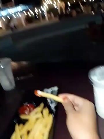 The video shows a close-up view of a table at night, with a person's hand dipping a french fry into ketchup. In the background, a street scene is visible, with lights from a building and passing vehicles. The setting appears to be an outdoor dining area. Two plastic cups with straws, a black pouch, and two smartphones are on the table. The time of day is clearly night due to the artificial lighting and the dark sky. The weather is not discernible from the video. The location is implied to be Uleeue, Indonesia, based on the context provided. The person's hand is holding the french fry, suggesting they are about to eat. The overall mood seems casual and relaxed, likely a moment of enjoying a meal or snack. No text is visible in the video.