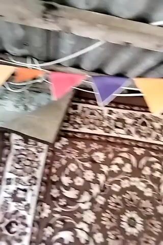 This video shows a close-up view of a room, likely an attic or an unfinished space, with a corrugated roof and wooden beams overhead. A string of colorful triangular pennants, in shades of red, purple, orange, and green, is strung across the ceiling. Below the pennants, a large, ornate rug with a floral pattern is hanging or draped. In the center of the frame, a bright LED lightbulb is suspended from a wire, illuminating the scene. The video pans slightly, offering glimpses of other parts of the room and the continuation of the pennant string. The lighting suggests it might be daytime, but the overall impression is of a rustic or makeshift setting.