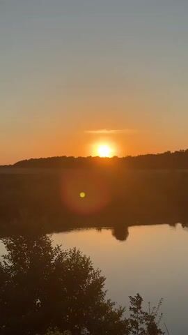A vertical video captures a sunset over a body of water, possibly a river. The sun is low on the horizon, casting a warm orange glow across the sky and reflecting on the water. A silhouetted treeline forms the backdrop. In the foreground, the tops of trees are visible. A circular lens flare appears in the middle of the frame. The sky transitions from a pale blue at the top to a vibrant orange near the horizon. The water is calm and reflects the sky's colors. The overall impression is of a serene natural landscape at dusk.