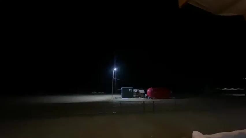 A night-time video shows a dark landscape with a few illuminated objects. In the center of the frame, a tall pole with a light shines downwards onto a sandy area. To the right of the pole, a small, dark rectangular structure is visible. Further to the right, a bright red, rounded object, possibly a trailer or a small vehicle, is illuminated by the pole light. In the foreground, a portion of a wooden ceiling with beams is visible, and the bottom of the frame shows some indistinct shapes that appear to be a railing or furniture. The overall scene suggests an outdoor, possibly remote location at night.