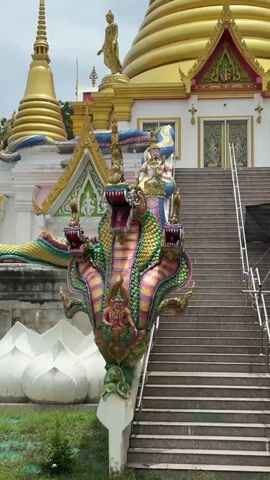 A statue of a multi-headed serpent with a human-like figure riding on its neck stands in front of a temple. The serpent is ornately decorated in vibrant colors, with green, yellow, pink, and blue scales. The human figure is adorned in traditional attire and holds a staff. Behind the statue, a wide staircase leads up to the temple entrance. The temple itself features a golden dome and intricate architectural details. The sky above is overcast.