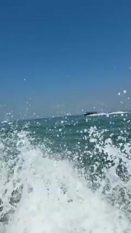 The video shows a speedboat moving quickly through blue water under a clear blue sky. The camera appears to be on a second boat that is splashing through the water. The speedboat is leaving a white wake behind it.