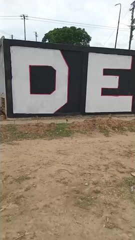 The video pans across a wall with large, bold lettering painted on it. The letters appear to spell out "DEPOR VIDA" with some additional text or imagery that is partially obscured. The lettering is in white with red and black accents, and some sections of the wall are painted red or black. In front of the wall is a dirt path, with some scattered debris, including plastic bottles and paper. Beyond the wall, a chain-link fence encloses a sports field or court. The sky is overcast, and there are trees and utility poles visible in the background. The overall scene appears to be an outdoor public space, possibly a sports complex or recreational area.