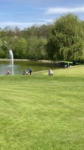The video captures a serene and sunny day on a golf course in Bad Rappenau, Germany. The scene is dominated by a large, well-maintained green lawn in the foreground and mid-ground, leading to a substantial pond. A prominent white water fountain continuously sprays water in the center of the pond, creating a focal point.

Dense green trees and foliage line the far bank of the pond, suggesting a natural, wooded boundary to the course. The sky above is clear and blue with only a few scattered white clouds, indicating pleasant weather conditions.

Several golf push carts are visible along the edge of the pond, suggesting ongoing play. At least two individuals are present: one person wearing a blue shirt is seen walking near a golf cart by the water, while another person in a green shirt is visible further to the right, also near a cart. Their presence and the equipment indicate that golfing activities are taking place.

The video slowly pans from left to right, offering a wider view of the picturesque landscape, encompassing the pond, fountain, green fairways, and surrounding trees. The overall impression is one of a peaceful and recreational outdoor environment on a bright, beautiful day. No discernible text is visible in the media.