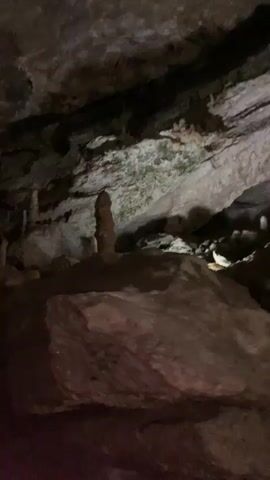 The video shows a group of people exploring a cave. The cave has rocky walls and ceilings, with stalactites hanging down in places. The people are walking through the cave, looking at the formations and taking pictures with their phones. The lighting in the cave is dim.