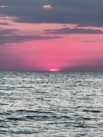 This is a vertical video showcasing a sunset over a body of water. The sun, a vibrant pink hue, is partially submerged on the horizon, casting a dramatic glow across the sky. The sky is filled with scattered clouds, tinged with shades of pink and purple, transitioning to a darker gray towards the top of the frame. The water is choppy, reflecting the colors of the sunset and creating a textured surface. A faint line of buoys or markers is visible across the water in the middle ground. The scene evokes a sense of tranquility and natural beauty. The time of day is clearly sunset due to the low position of the sun and the colorful sky. There is no visible text in the video. The location context provided is Челядиновское сельское поселение, Ukraine, suggesting the scene takes place in this region of Ukraine.