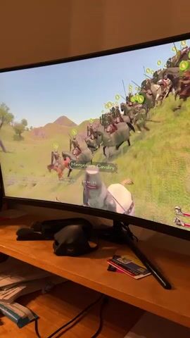 The video shows a computer monitor displaying a video game. The game appears to be a medieval battle simulation, with knights on horseback engaged in combat. In the foreground of the game, a knight in silver armor is on a white horse, holding a bow. In the background, a large group of mounted soldiers, identified as "Mercenary Cavalry," are charging uphill. The monitor is on a wooden desk, and in front of the monitor, there is a black mask and some papers. The lighting suggests an indoor setting at night.