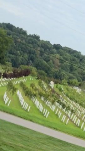 The video shows a view of a cemetery from a moving vehicle. The cemetery has rows of white markers or headstones on a green, grassy hillside. There are trees scattered throughout the cemetery, and a darker line of trees or foliage forms the backdrop. A paved road or path runs horizontally across the lower portion of the frame, separating the foreground grass from the cemetery. The sky is visible above the trees, with some clouds. The footage appears to be shaky, suggesting it was recorded handheld while in motion.
