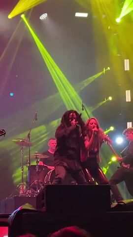 A concert performance is underway, illuminated by vibrant green and yellow stage lighting that casts dramatic beams across the smoky atmosphere. In the foreground, a band performs on a raised stage. A male vocalist, distinguished by his dreadlocks and dark clothing, is front and center, energetically singing into a microphone and gesturing with his hands. To his right, a female vocalist with blonde hair, also in dark attire, stands close by, holding a microphone and singing. Behind them, several other band members are visible: a guitarist on the far right playing a red electric guitar, a drummer on the left behind a drum set, and another musician in the center background. The band is positioned on a dark stage, with what appear to be amplifiers and speakers in the immediate foreground. In the lower portion of the frame, audience members' hands and cameras are visible, indicating the perspective is from within the crowd.