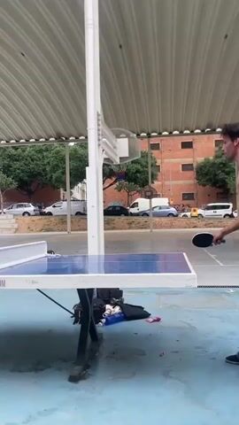 A young man in a white t-shirt and jeans is playing table tennis. He is holding a red table tennis paddle and is about to hit the ball. The table tennis table is blue and white and is outdoors. There is a white net in the middle of the table. In the background, there are trees and buildings. There is also a basketball hoop. Another young man in a black t-shirt is also present, but not actively playing at this moment. The ground is blue and wet, suggesting it may have rained. The video is shot from a low angle, close to the table.