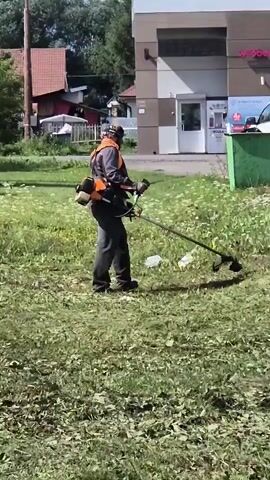 A person wearing a grey jacket, orange vest, dark pants, black shoes, and a camouflage hat with goggles is using a string trimmer to cut grass. The person is walking from left to right across the frame, swinging the trimmer in front of them. The grass is green and tall in some areas, with cut grass visible on the ground. In the background, there is a light-colored building with a glass entrance and a green dumpster. There are also cars parked in front of the building and some houses in the distance. The scene appears to be an outdoor public space.