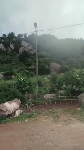 The video shows a mountain scene on a cloudy day. Initially, there is a view of a tree-covered hillside with a light pole. Two people wearing green vests are working near the bottom of the slope. The camera then pans to show a dirt road leading up the mountain, with a white building visible on the slope. The camera continues to pan, revealing a more distant view of white buildings nestled among the trees on the mountainside, before shifting to a broader view of a city in the distance, also with white buildings. The sky is overcast with dark clouds, but some patches of blue sky can be seen.