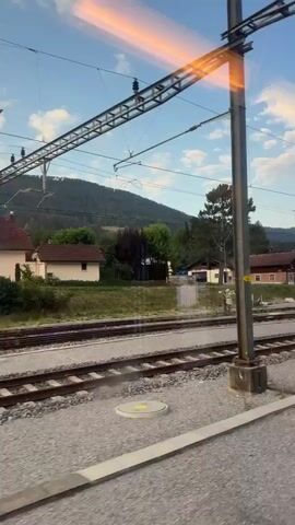 This is a perspective shot from a moving train looking out a window. The train is on tracks in a rural or suburban area. The immediate foreground shows the gravel bed and rails of a train track, as well as a blurry white barrier or object in the lower right. In the middle ground, a second set of train tracks is visible, with a grassy embankment beyond. Several houses and buildings are situated on the embankment, some with dark-colored roofs, others with lighter-colored facades. A large tree stands prominently among the buildings. The background consists of a tree-covered hillside under a blue sky with scattered white clouds. A distinct horizontal, blurry band of orange and yellow light is visible in the sky, suggesting either a reflection on the window or a sunset. Power lines and poles related to the train's operation are visible throughout the scene. There are no people clearly visible or interacting in the shot.