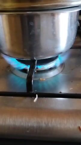 A stainless steel pot sits on a gas stove burner, with a blue flame visible beneath the pot.  A stainless steel lid is on the pot. The burner is part of a stainless steel stovetop.  A piece of uncooked rice lies on the stovetop near the burner.  The audio features the distinct sound of a baby crying.