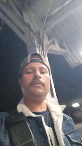 A man films himself in selfie mode while walking on a train platform at night. He is wearing a trucker cap, a denim jacket with a white fleece collar, and a dark t-shirt.  He has a mustache and a bag slung over his shoulder. The platform is illuminated by overhead lights. In the background, a train car can be seen.  The camera is shaky, indicating the man is moving.