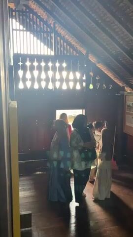 A group of people are gathered inside a wooden building with a sloped, beamed ceiling. Sunlight streams through decorative openings in the upper wall, casting a patterned light onto the wooden floor and walls. Most individuals are wearing modest clothing, including headscarves. Some are looking at framed pictures on the wall, while others appear to be conversing or moving through the space. The atmosphere seems to be one of a tour or a visit to a historical or cultural site.