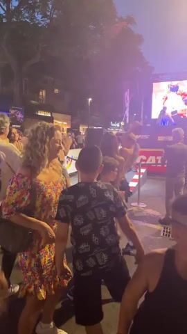 This is a video of an outdoor event at night. In the foreground, several people are visible, some with their backs to the camera. In the middle ground, there is a stage with a large screen displaying images or video. A DJ is visible on the stage, and a crowd is gathered in front of it. The background shows buildings and trees, and the sky is dark with some streetlights visible. The overall atmosphere appears to be festive, with people dancing and enjoying the event. The video is grainy and the lighting is dim, suggesting it was shot with a mobile phone camera in low-light conditions.